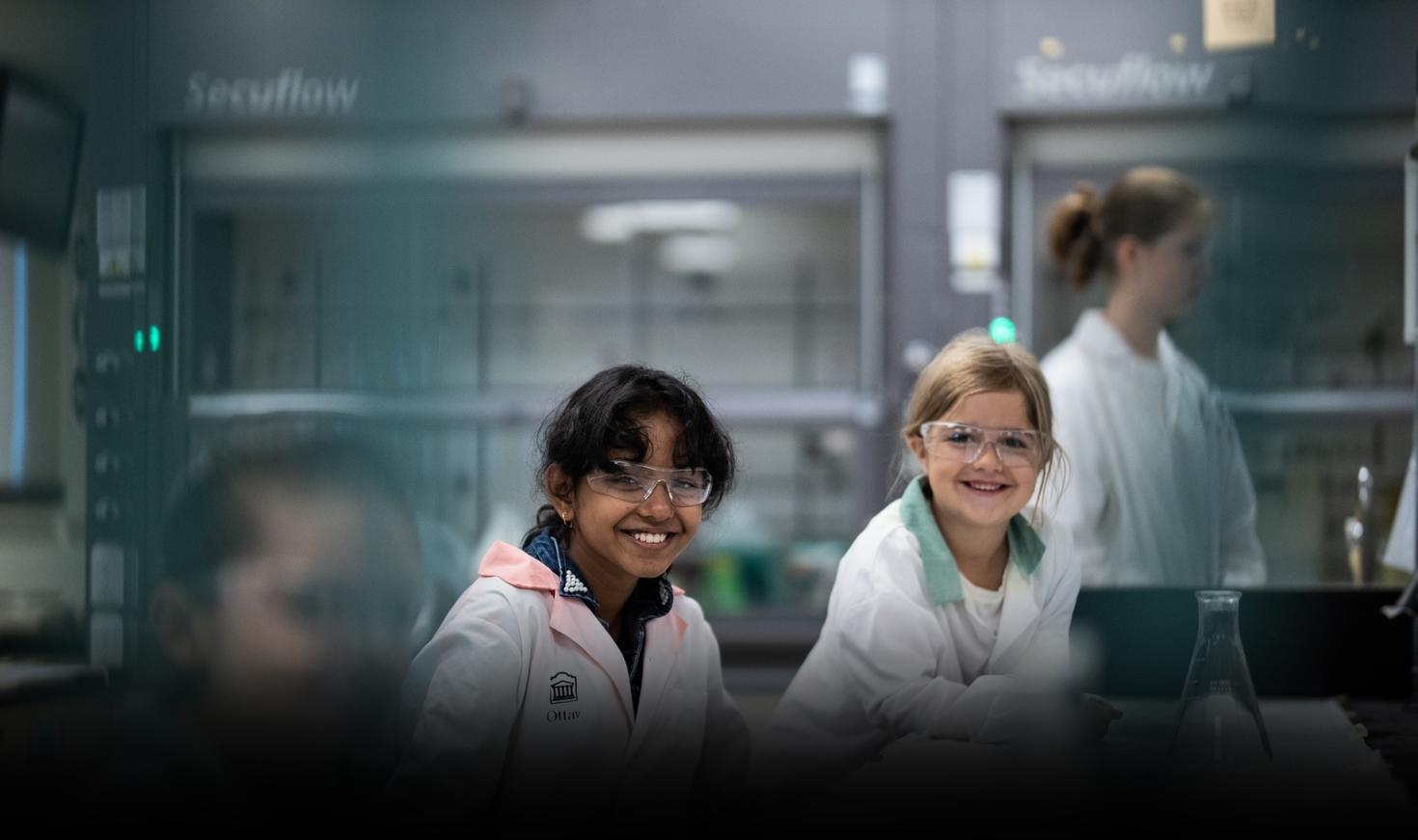 Two kids smiling in a lab wearing lab coats and protective eyewear
